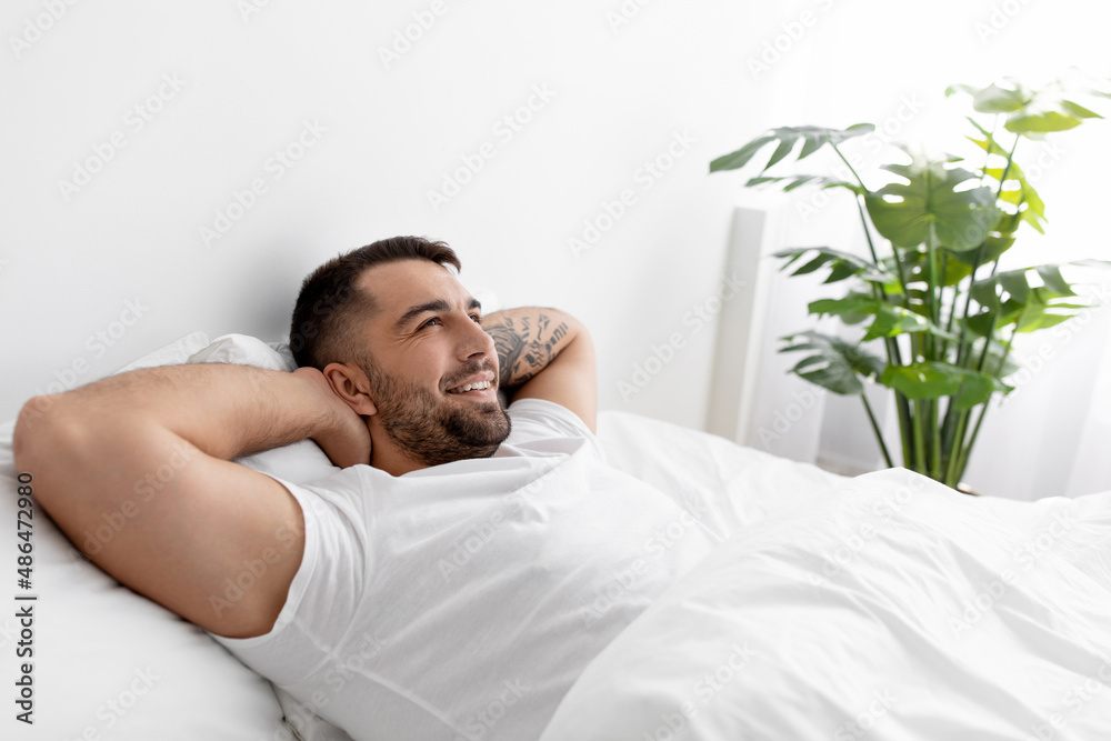 Happy caucasian young male in white t-shirt rests and relaxes, looks at empty space on bed in bedroom interior Happy caucasian young male in white t-shirt rests and relaxes, looks at empty space on bed in bedroom interior