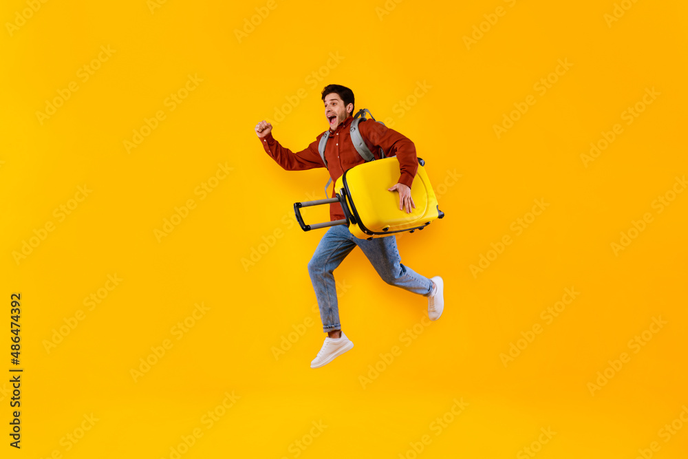 Emotional Male Tourist Jumping Carrying Suitcase Over Yellow Studio Background