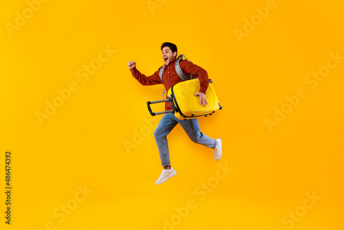 Emotional Male Tourist Jumping Carrying Suitcase Over Yellow Studio Background