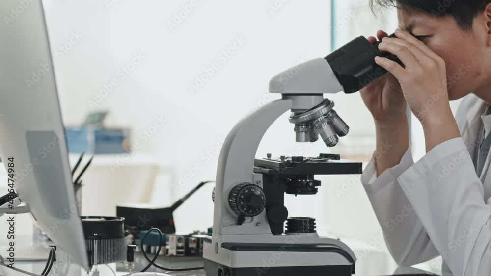 Tracking shot of Asian female engineer in white coat looking through microscope while doing research in laboratory