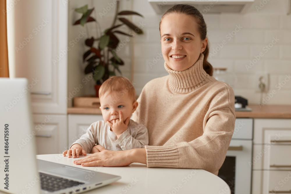 Portrait of attractive young adult woman wearing beige sweater sitting in kitchen and working online on laptop, holding baby, looking at camera with charming smile. Portrait of attractive young adult woman wearing beige sweater sitting in kitchen and working online on laptop, holding baby, looking at camera with charming smile.