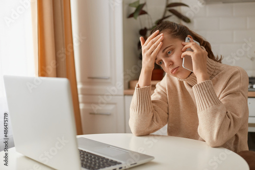 Horizontal shot of tired or sick woman wearing beige sweater sitting in kitchen and working online on laptop and talking on smartphone, having problems or suffering headache.