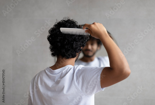 Back view of indian curly man combing hair standing near mirror in bathroom. Male haircare routine concept photo