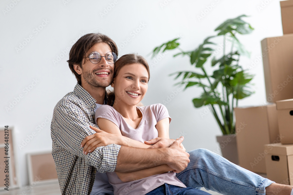 Smiling millennial european husband in glasses hugs wife in room interior with cardboard boxes with stuff