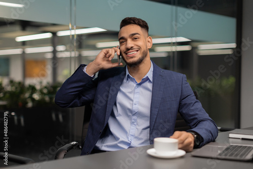 Canvas Cheerful arab entrepreneur sitting at worktable, talking on phone