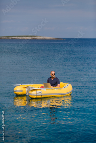Digitaler Nomade mit Sonnenbrille sitzt in gelbem Gummiboot am Meer und arbeitet am Laptop, Hochformat