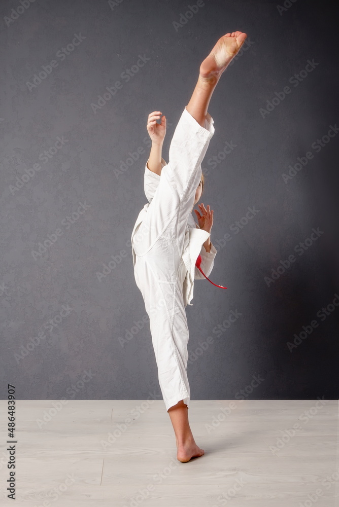 young girl karateka in a white kimono and a red belt trains and performs a set of exercises against a gray wall