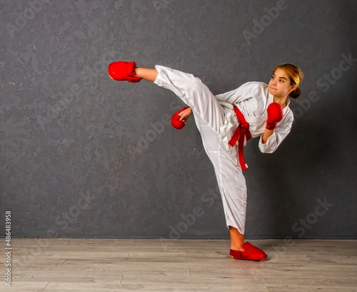 young karateka girl in a white kimono and red competition outfit trains and performs a set of exercises against a gray wall