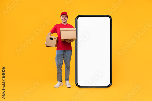 Courier Guy Near Huge Phone Screen Holding Boxes, Yellow Background © Prostock-studio