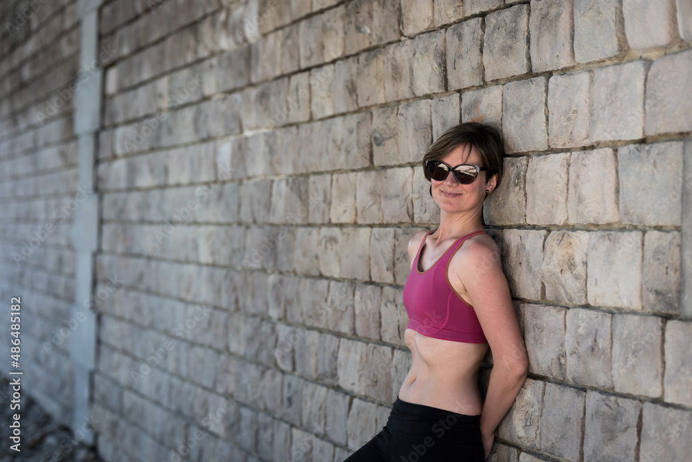 Young white smiling woman in sports bra posing against a rough wall.