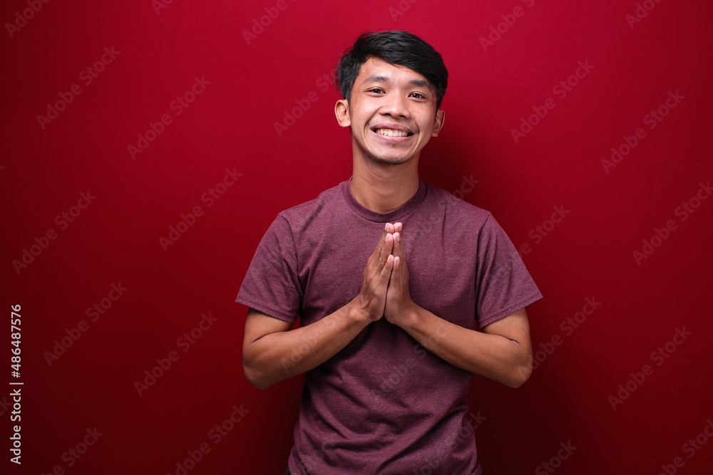 Asian youth wishful expression, making a prayer and wishing gesture over a red background Asian youth wishful expression, making a prayer and wishing gesture over a red background