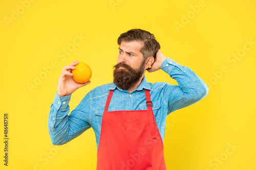 Puzzled man in apron looking at fresh orange fruit scratching head yellow background, fruiterer
