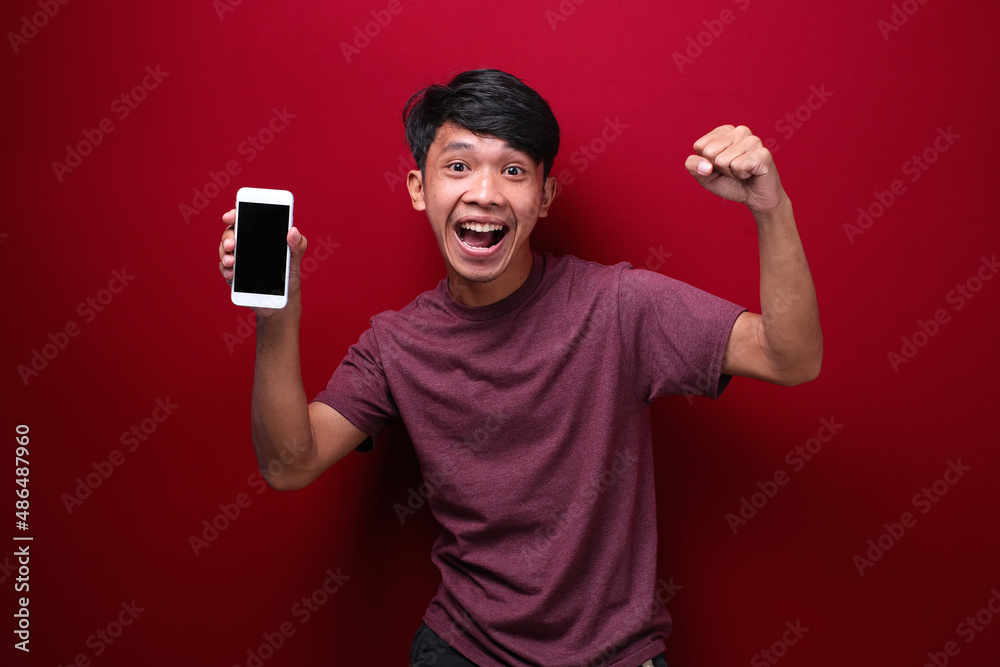 happy young asian guy smiling to camera while holding smartphone pointing at cell phone screen happy young asian guy smiling to camera while holding smartphone pointing at cell phone screen