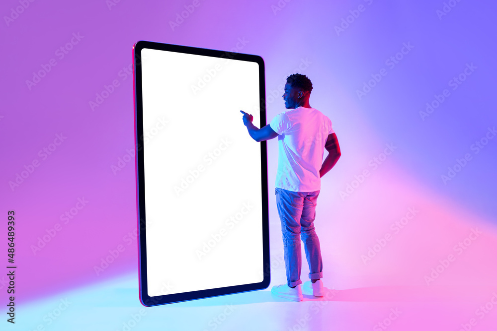 Happy young African American man standing near big tablet computer with mockup, touching screen in neon light Happy young African American man standing near big tablet computer with mockup, touching screen in neon light