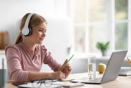 Smiling pretty european teenager girl student in headphones watches video lesson at table in kitchen