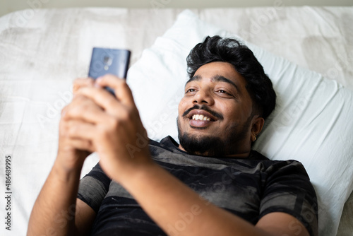 happy young man chatting on mobile phone while sleeping on bed - concept of using social media,dating app, flirting and technology. photo