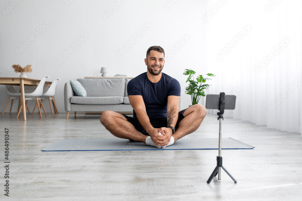Fototapeta premium Happy european millennial guy sitting on mat looking at smartphone webcam recording sports blog in living room