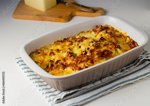 Potato casserole with cheese in ceramic form on a napkin on a white table. Nearby is a cutting board with a piece of cheese and a knife.