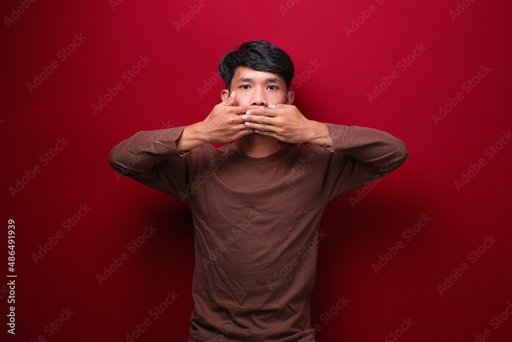 asian man on red background wearing brown t-shirt covering mouth with hand asian man on red background wearing brown t-shirt covering mouth with hand