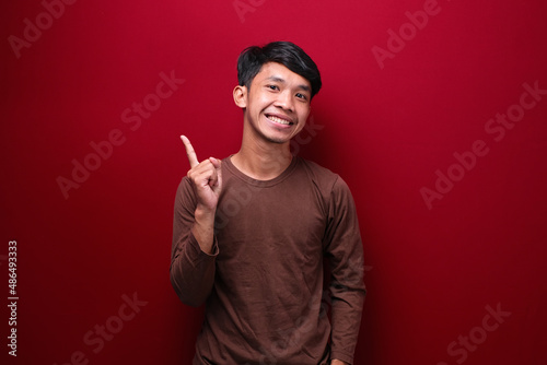 asian man on red background wearing brown t-shirt pointing to empty space