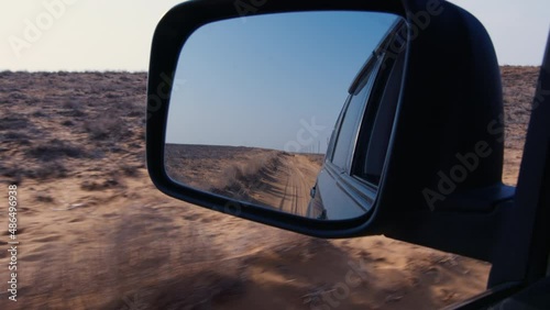View of side mirror of car, which reflects unpaved road behind vehicle, dry plants, sky, electric power transmission poles. Sunny summer day in barren area. Auto driving on soil route in desert place photo