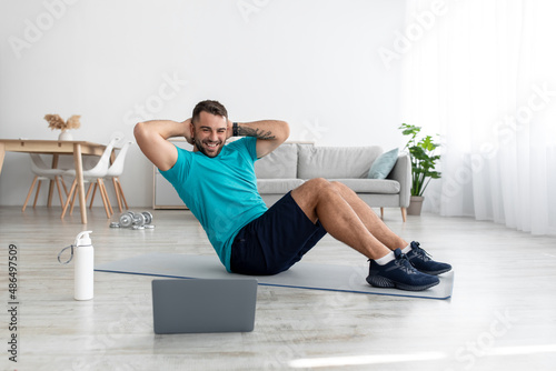 Cheerful young muscular caucasian guy doing abs exercises and looking at computer in living room interior