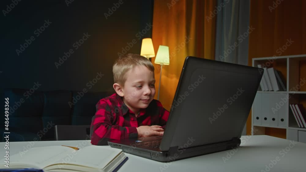 The child teaches lessons while at home sitting in the living room at the table using a laptop, books, notebook and looking at the camera and smiling. The child teaches lessons while at home sitting in the living room at the table using a laptop, books, notebook and looking at the camera and smiling.