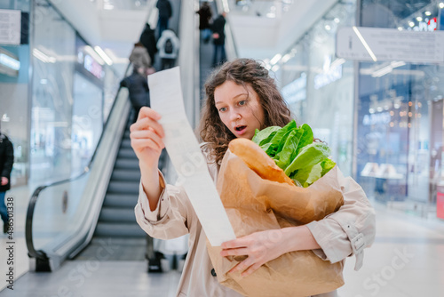 A woman in shock looks into a paper check from a supermarket in a shopping center against the background of an escalator and holds a package with fresh products, price increase photo