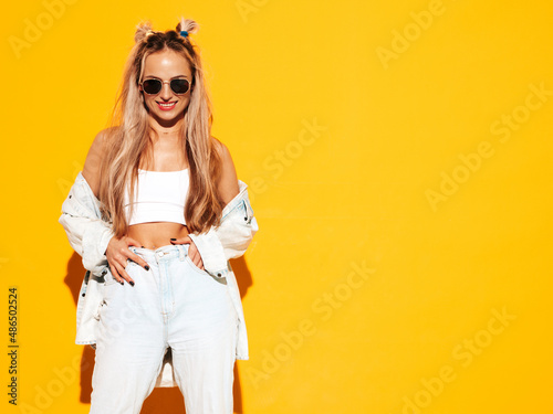Portrait of young beautiful smiling blond female in trendy summer clothes. Sexy carefree woman posing near yellow wall in studio. Positive model having fun indoors. Cheerful and happy in sunglasses © halayalex