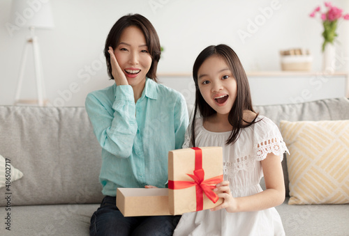 Excited mature Asian woman and her adorable little granddaughter opening gift on couch at home