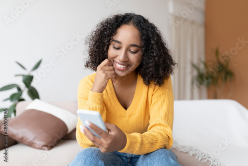 Young black woman using cellphone, having online video call, sitting on sofa, communicating in social network at home