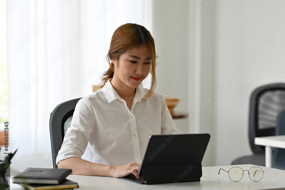 A portrait of a young pretty Asian businesswoman in a white shirt working on a touchpad tablet in the office. Business and work concept A portrait of a young pretty Asian businesswoman in a white shirt working on a touchpad tablet in the office. Business and work concept