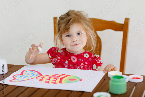 Little creative toddler girl painting with finger colors a fish. Active child having fun with drawing at home, in kindergaten or preschool. Education and distance learning for kids. Creaitve activity. photo