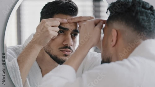 Unhappy arabian indian arab bearded man looking in bathroom mirror feeling worried about facial skin problem acne. Anxious millennial guy squeezing pimple touching face frustrated blackheads skincare