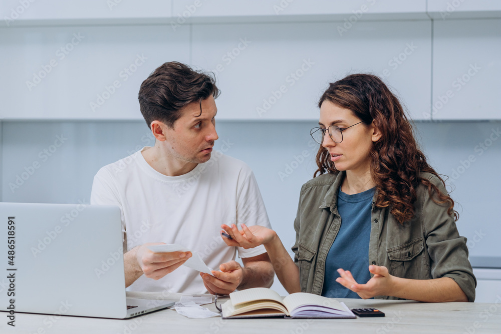Fototapeta premium Husband and wife calculate the expense of finances sitting at home in the kitchen at the table and write it down in a notebook, a woman with checks in her hands makes excuses, a man gets upset