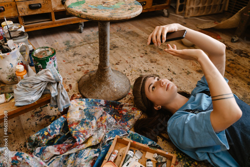 Young woman artist using mobile phone while lying on floor in her studio