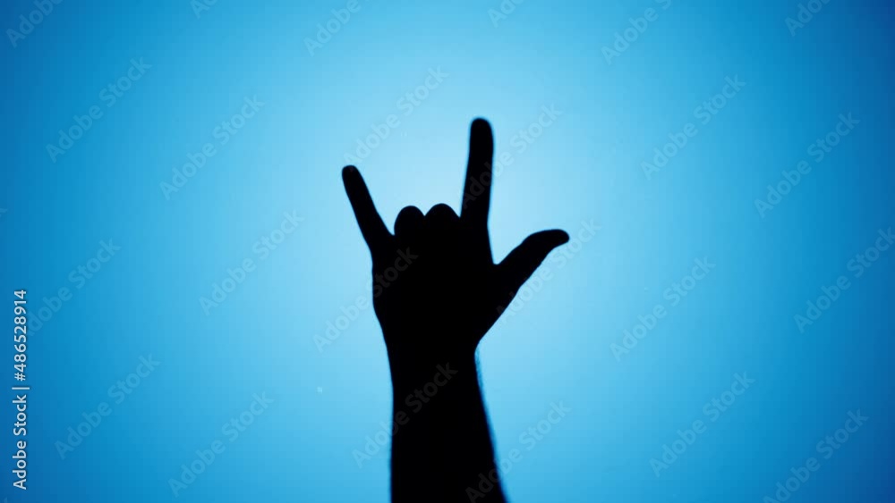 Man showing rock and roll gesture with fingers isolated on blue background. Male person making shadow silhouette with hand close-up. Man showing rock and roll gesture with fingers isolated on blue background. Male person making shadow silhouette with hand close-up.