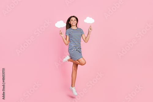 Photo of sweet shiny girl wear striped dress jumping high rising two clouds empty space isolated pink color background