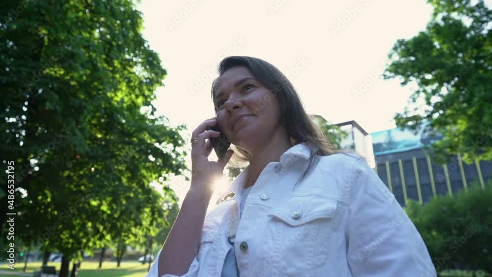Businesswoman in white stylish jacket stands talking on smartphone with partner against building in park slow motion close low angle shot Businesswoman in white stylish jacket stands talking on smartphone with partner against building in park slow motion close low angle shot
