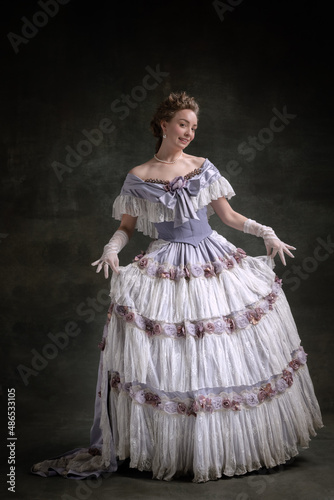 Full-length vintage portrait of young adorable girl in image of medieval royal person in renaissance style dress isolated on dark background. Comparison of eras