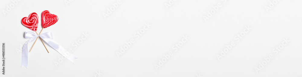 A long horizontal banner with two heart-shaped lollipops on a stick with a white bow on a white background. Valentine's Day greeting card. Copy space. Flat lay, top view. A long horizontal banner with two heart-shaped lollipops on a stick with a white bow on a white background. Valentine's Day greeting card. Copy space. Flat lay, top view.