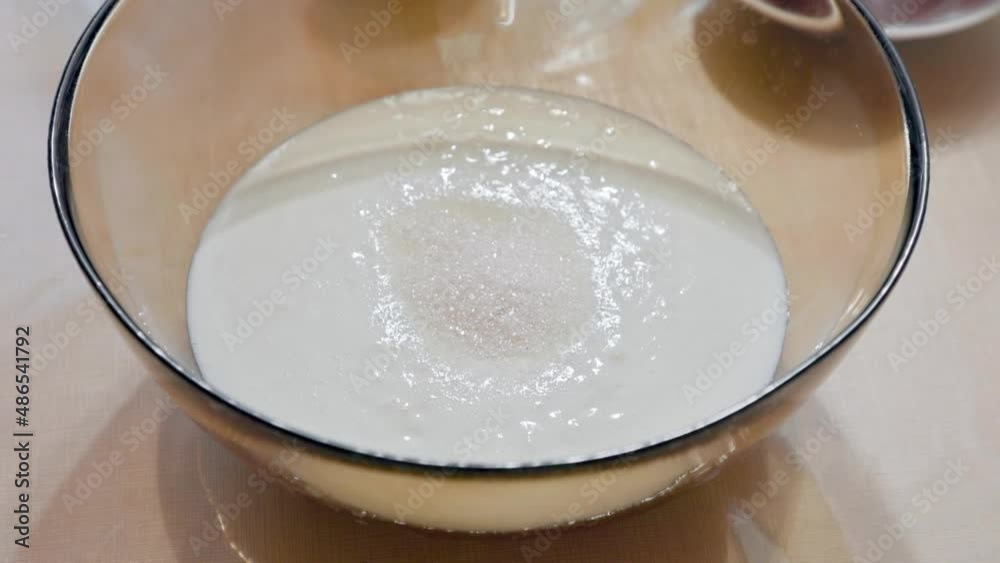 A plastic spoon of sugar is poured into a glass bowl with yogurt standing on the table. Close-up. Home cooking of food. A plastic spoon of sugar is poured into a glass bowl with yogurt standing on the table. Close-up. Home cooking of food.