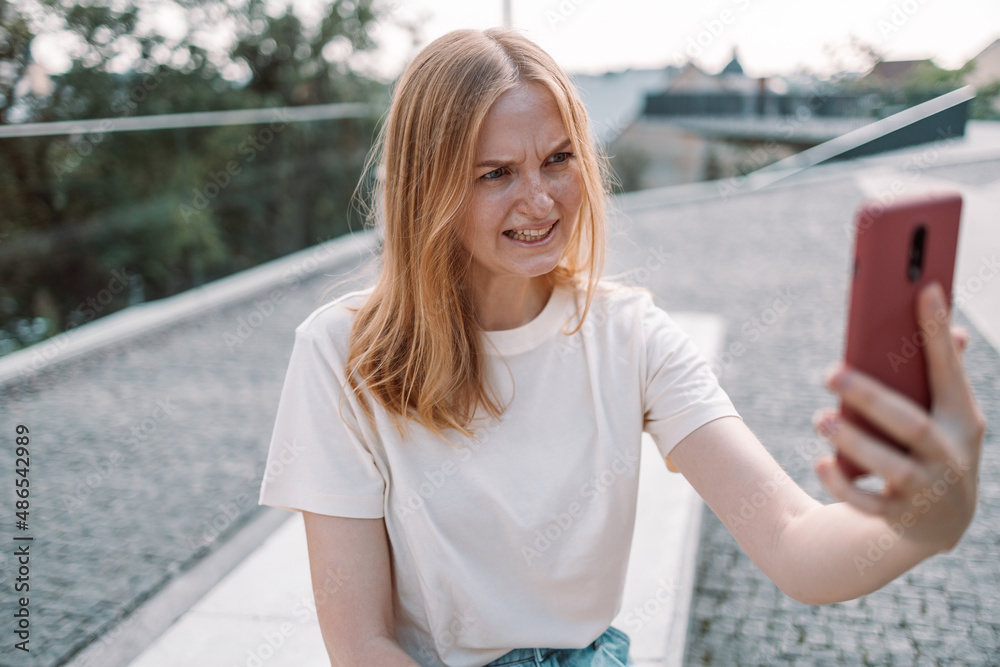 Blonde hair angry young woman posing outdoors in park take a selfie by mobile phone while standing in the park. Blonde hair angry young woman posing outdoors in park take a selfie by mobile phone while standing in the park.