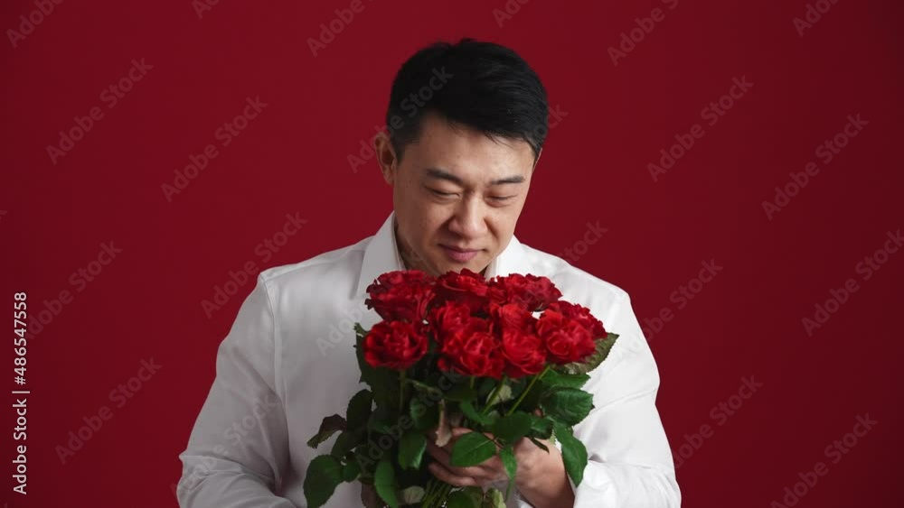 Positive Asian man in white shirt holds out a bouquet of red roses to the camera in the red studio Positive Asian man in white shirt holds out a bouquet of red roses to the camera in the red studio