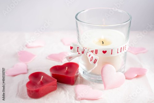 The theme is Valentine's Day. A burning candle and two red candles in the shape of hearts and decorative pink hearts are on a white background. Free space for text.