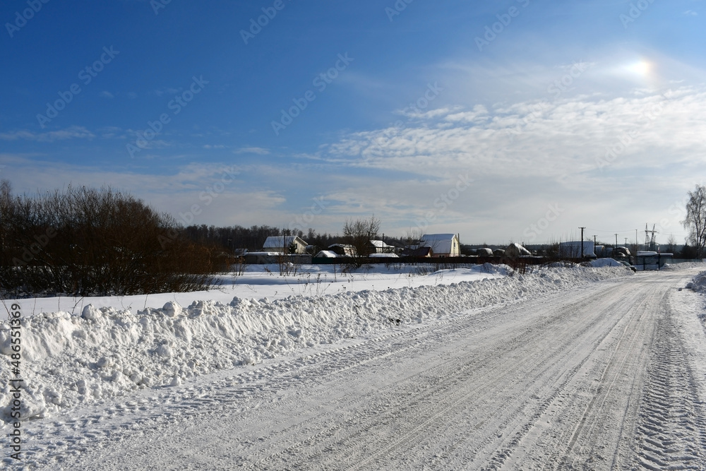 Fototapeta premium A frosty clear winter day. View of the snowy road leading to the village or country village, rural houses in the distance