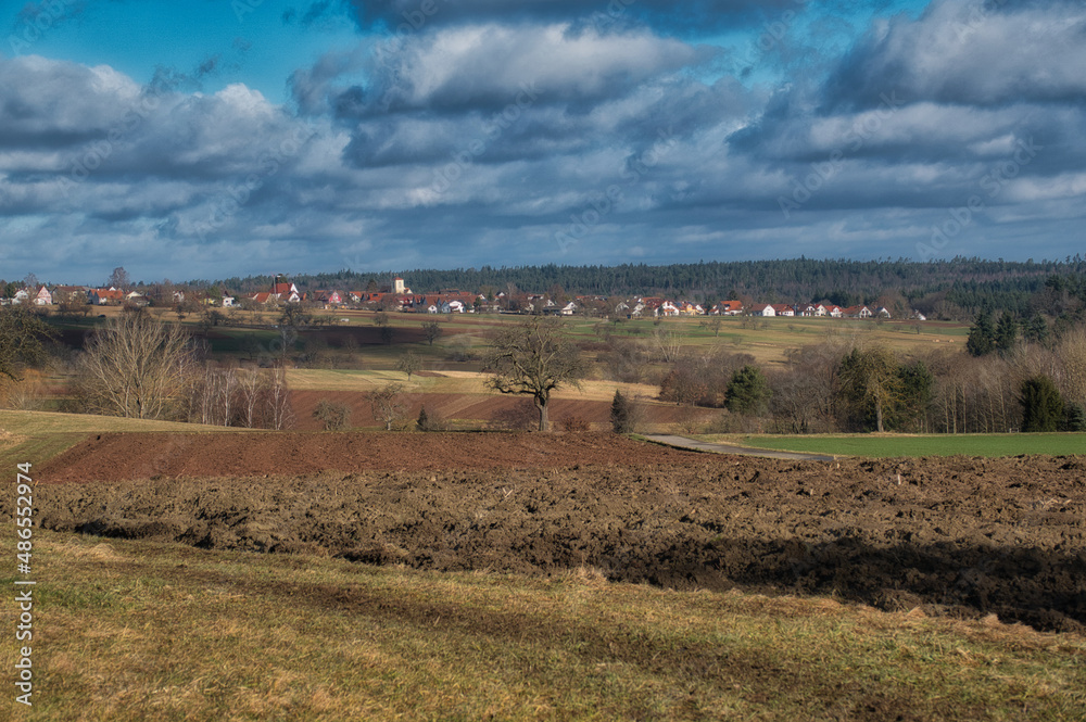 Ein umgepflügter Acker im Frühjahr mit einem Dorf im Hintergrund und kräftigen Wolken am Himmel Ein umgepflügter Acker im Frühjahr mit einem Dorf im Hintergrund und kräftigen Wolken am Himmel