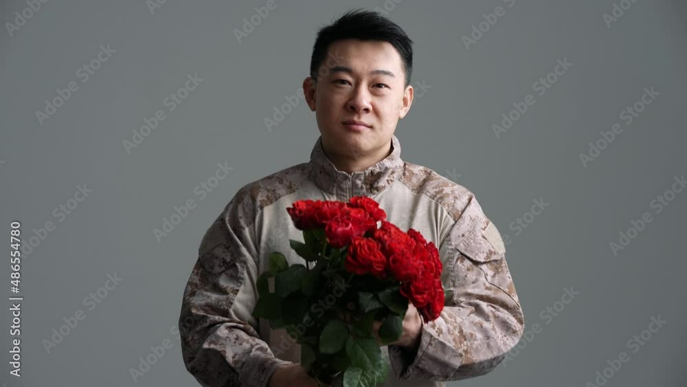 Smiling soldier Asian man holds out a bouquet of red roses to the camera in the grey studio Smiling soldier Asian man holds out a bouquet of red roses to the camera in the grey studio