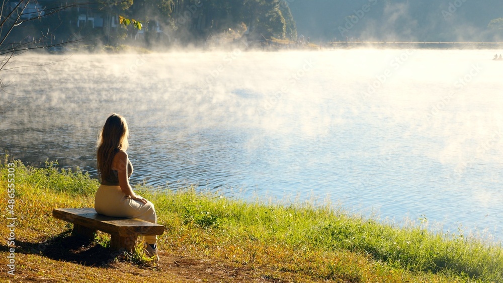 Travel woman relax on nature near misty lake. Female traveler sitting on bench, enjoy fog morning on river. Calmness, relaxation, meditation, freedom, beauty of nature concept