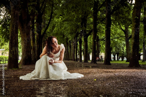 bride in white dress after rain completely wet on the ground on her knees between trees on a long path alley smiling praying shooting 